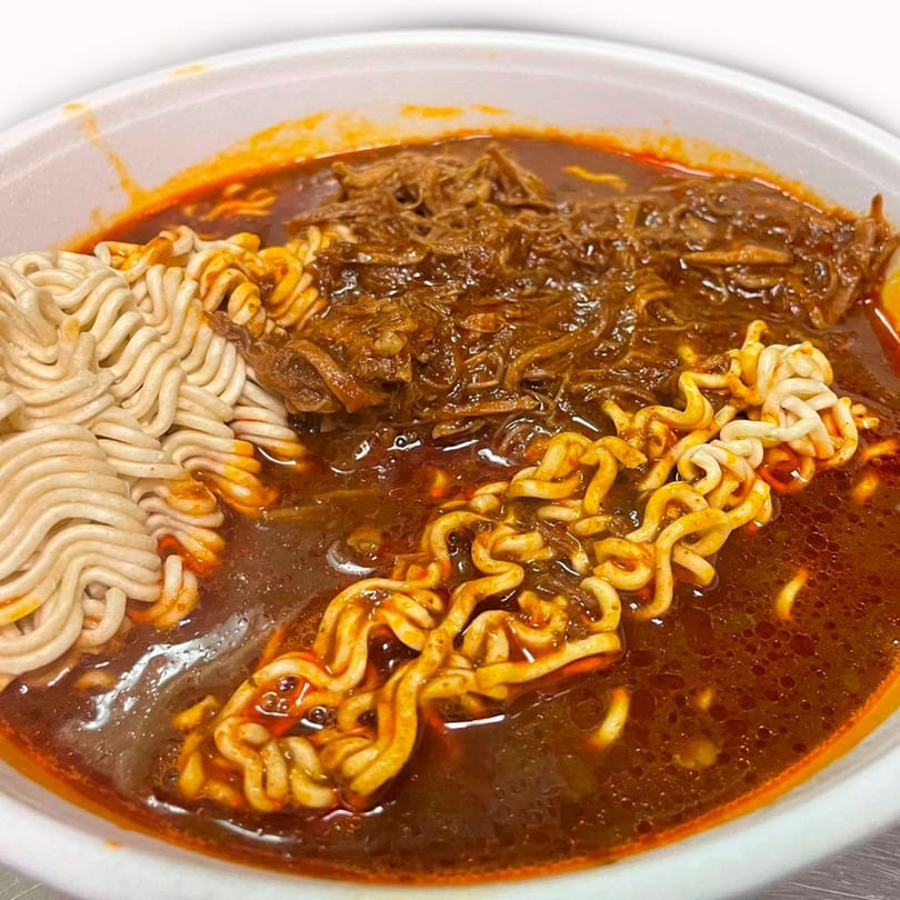 Close-up of a bowl of birria ramen with shredded meat and noodles in a rich red broth.