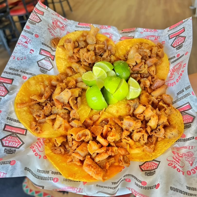 Four El Taco Loko tacos with crispy pork and lime wedges on branded paper.