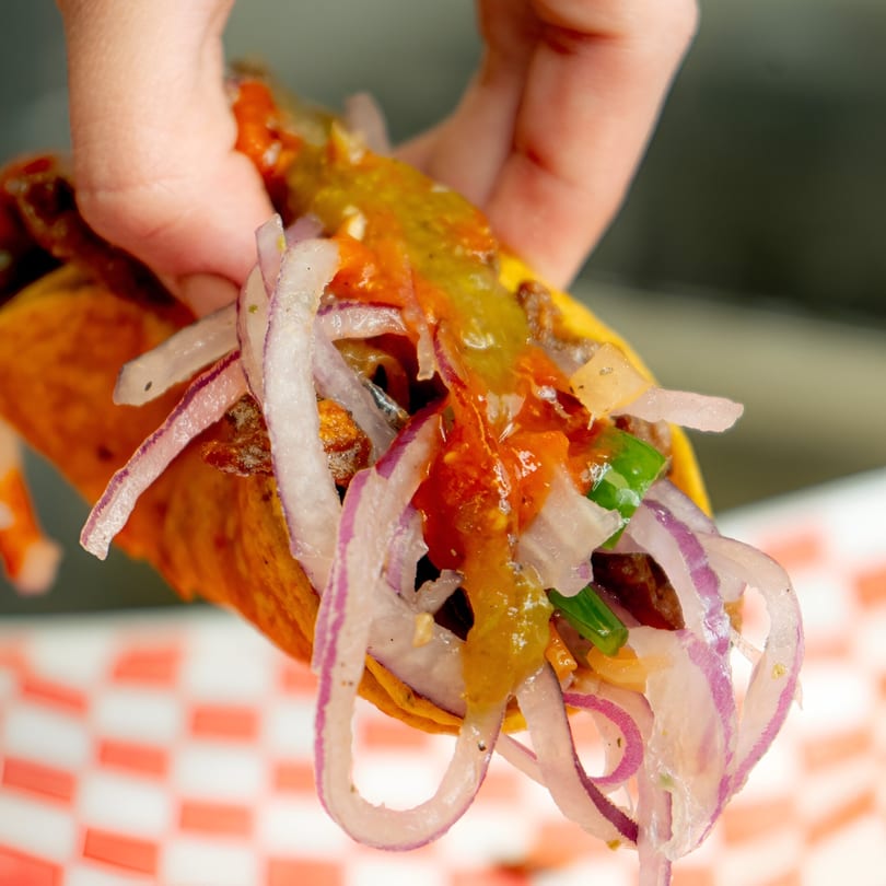 Hand holding a vibrant El Taco Loko taco filled with meat, red onions, and salsa.