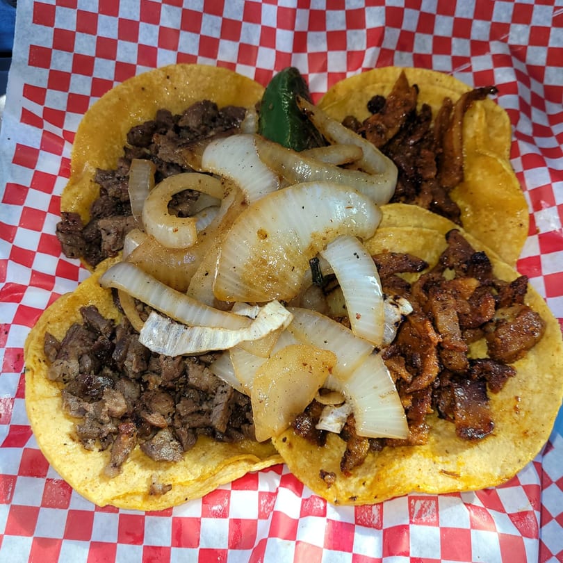 Four El Taco Loko tacos with meat and grilled onions on red and white checkered paper.